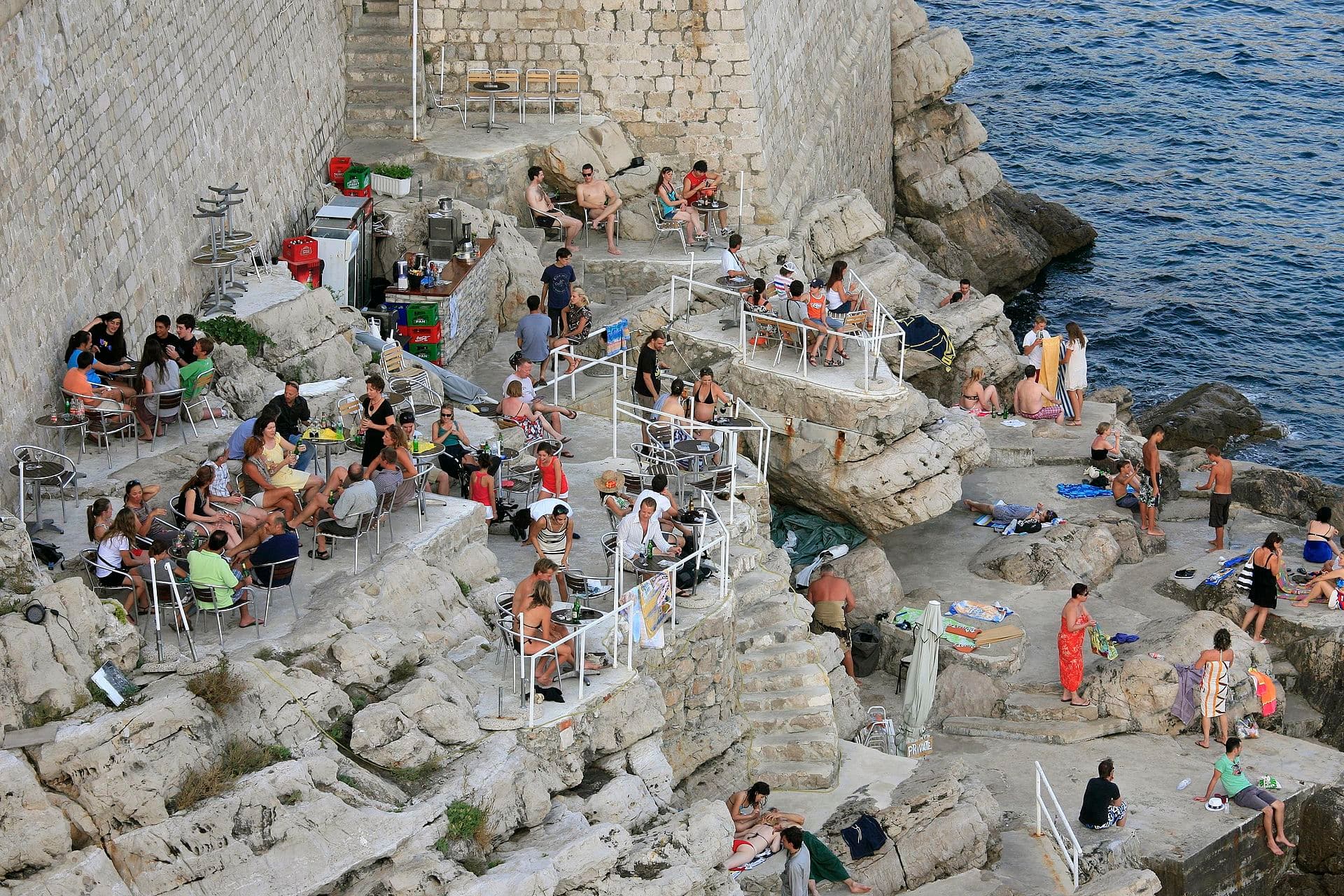 Una scala scavata nella roccia scende verso il mare proprio sotto le mura: qui, in un varco nascosto, si trova il Buža Bar, dove i locali sorseggiano un drink al tramonto lontano dalla folla di Stradun.